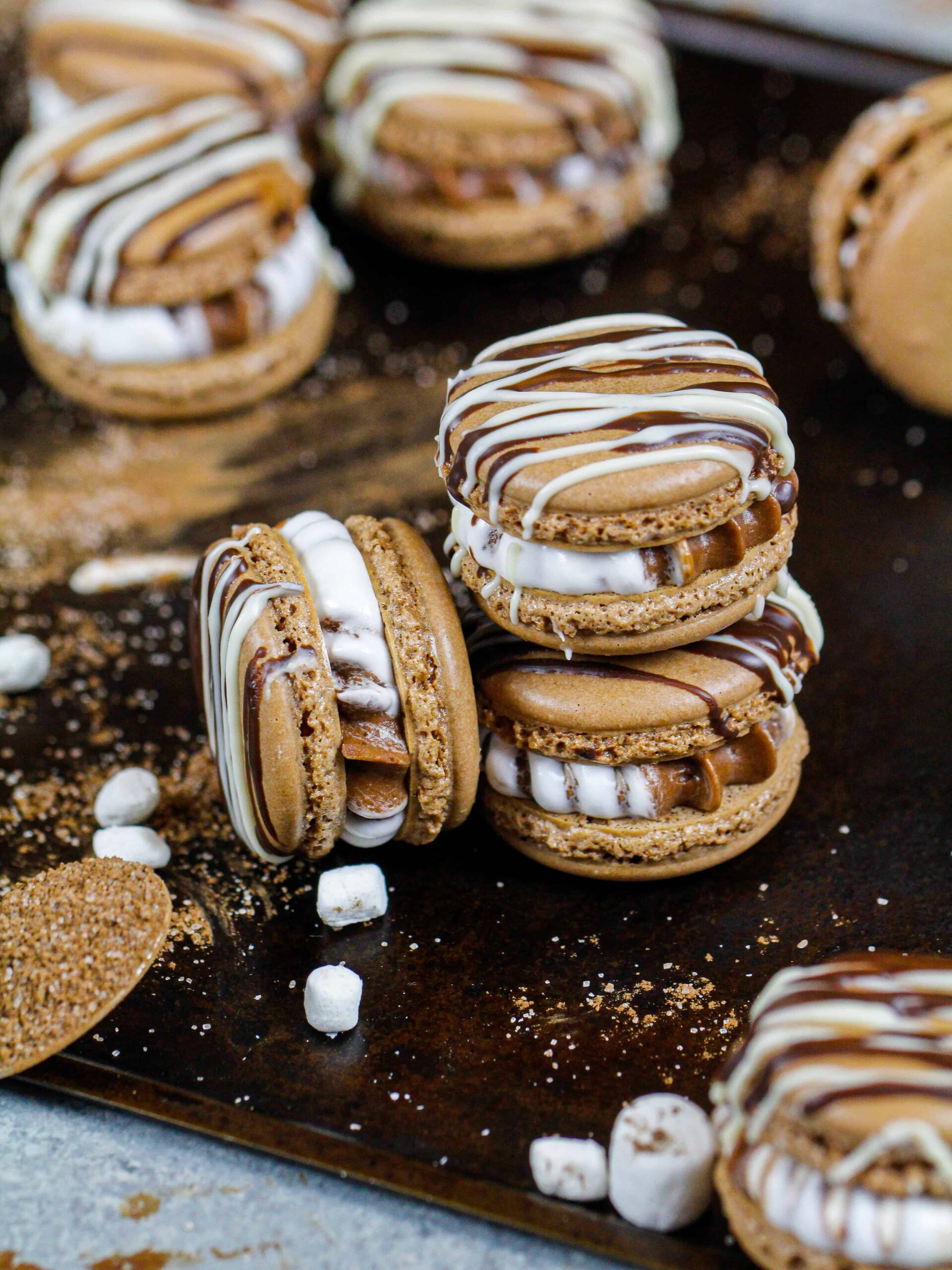 热可可马卡龙的图像，充满了热可可和棉花糖奶油奶油，它们被堆叠在一起的托盘包围着小棉花糖